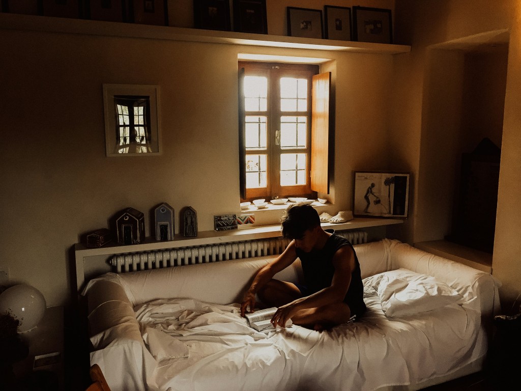 Picture of a lone young men sitting in his bed, reading a book with extreme focus. The dark and beige aesthetic of the small bedroom evokes a simplicity that illuminates the wanderlust of the young man, who is travelling far beyond the wal of his bedroom, through his book. 